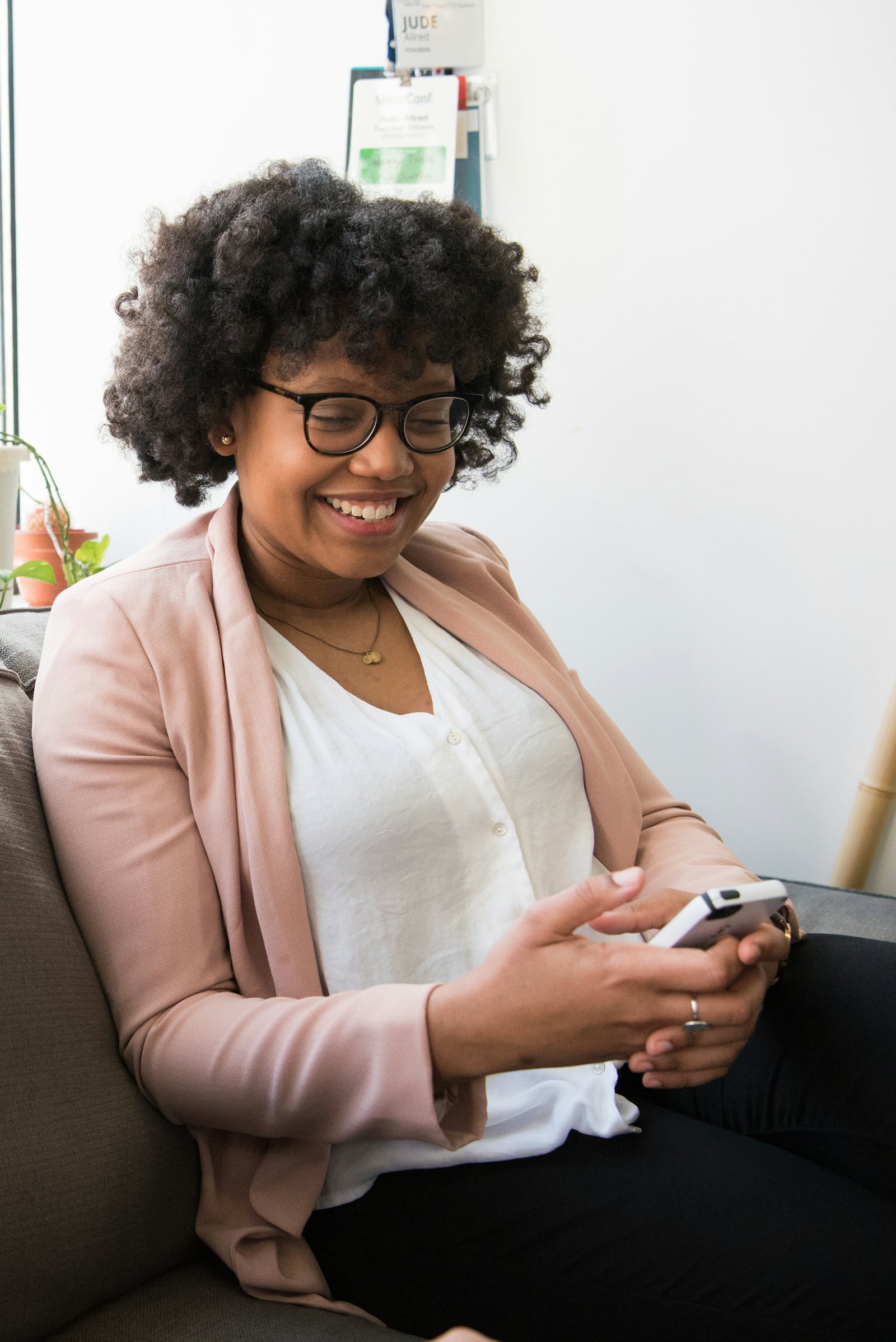 A smiling young Black woman in glasses checking her phone while seated on a sofa