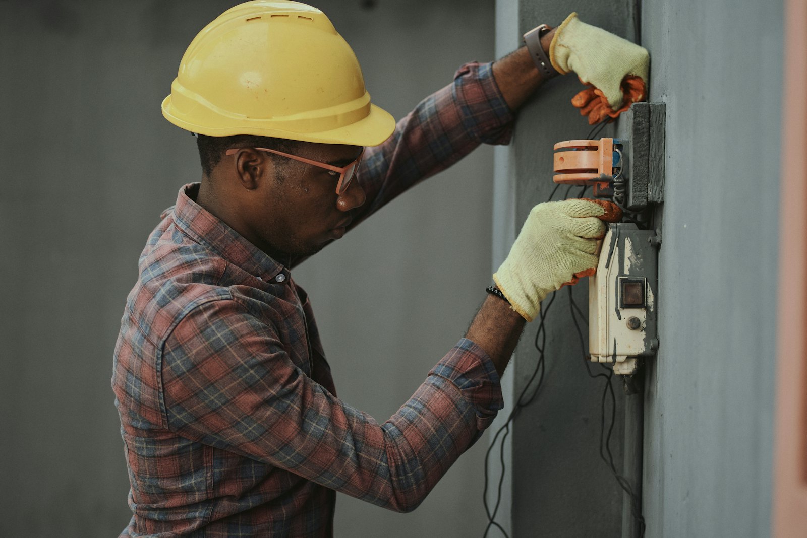 A Black technician in a yellow hard hat and gloves working on a meter box mounted on a wall